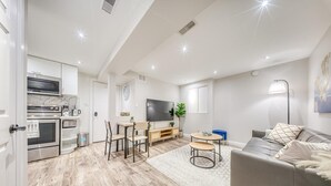 A modern living room with a grey sofa, a coffee table, and a TV. There is a kitchen area with a microwave, oven, and cabinets. The room has wooden flooring and a large window. - The Cozy Cove on Flatford (Mississauga)