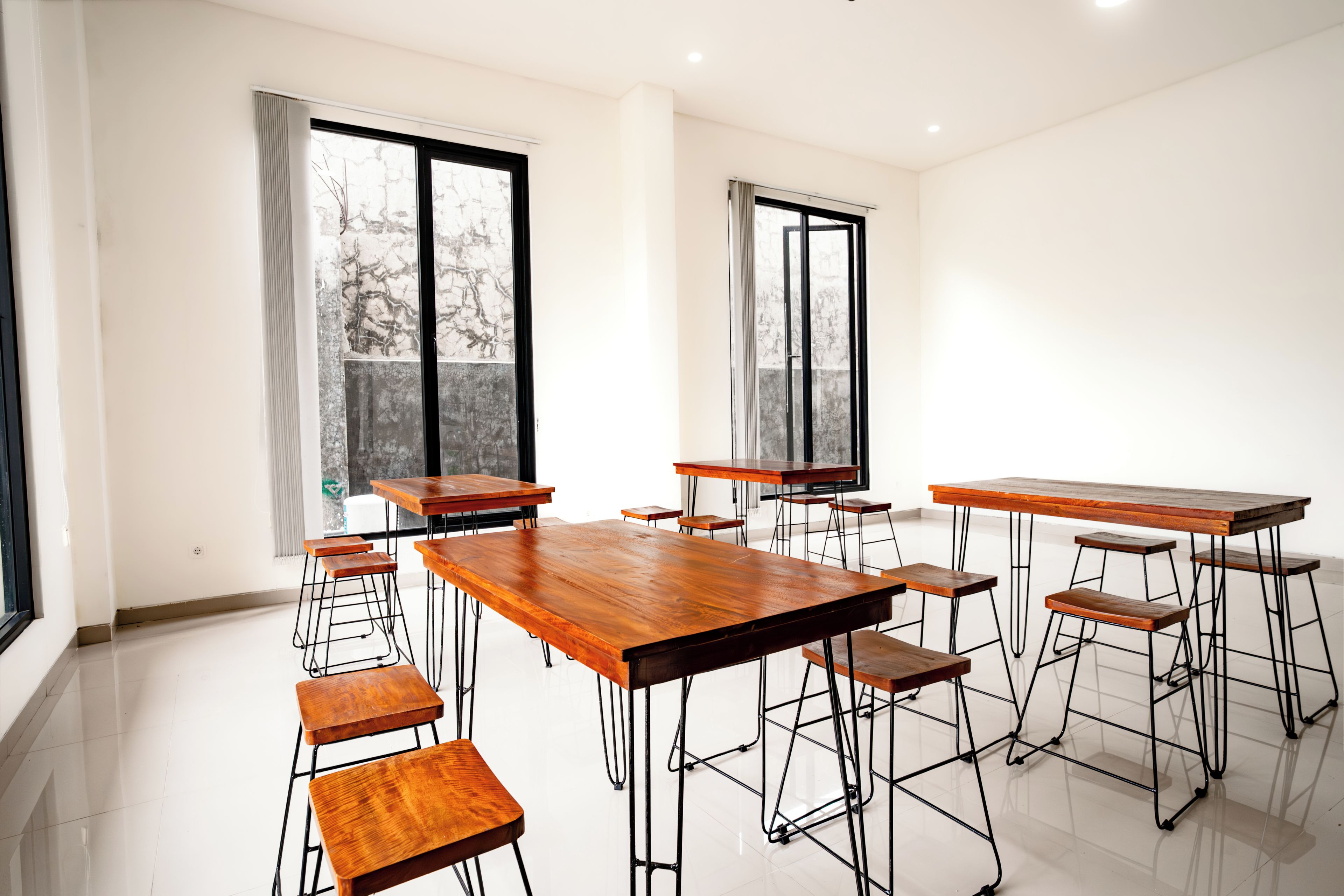 A spacious room with multiple wooden tables and stools, large windows with curtains, and a tiled floor.