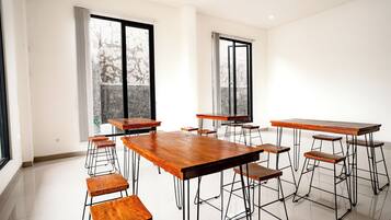 A spacious room with multiple wooden tables and stools, large windows with curtains, and a tiled floor.