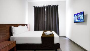 Habitación de hotel con una cama individual, una mesita de noche de madera, un televisor de pantalla plana en la pared y una ventana con cortinas oscuras.