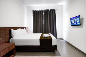 A hotel room with a single bed, a wooden nightstand, a flat-screen TV mounted on the wall, and a window with dark curtains.