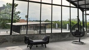Ein geräumiger Innenbereich mit hoher Decke und großen Fenstern, die einen Ausblick ins Freie bieten. Eine gepolsterte Bank und ein Hängekorbsessel sind vorhanden.