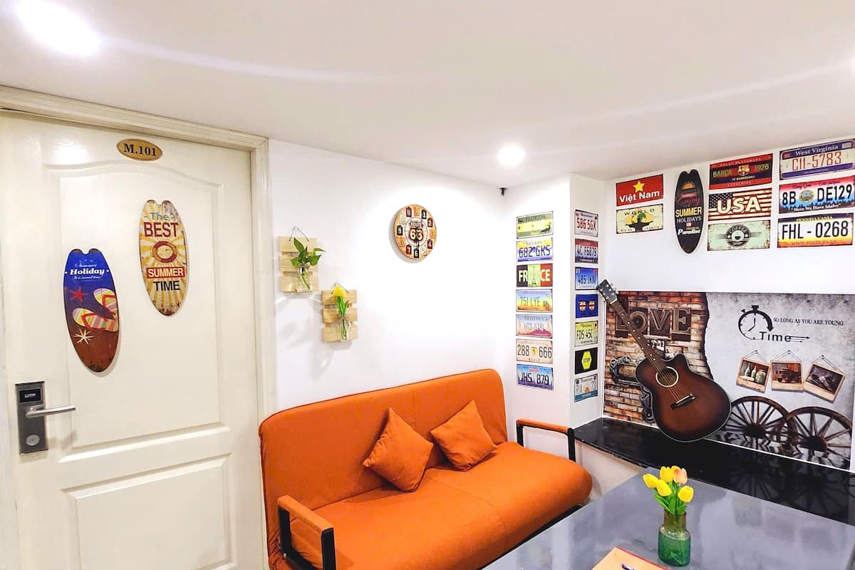 A room with a white door labeled "M.101," an orange sofa with two pillows, a small table with a vase of yellow flowers, and a wall decorated with various license plates and a guitar.