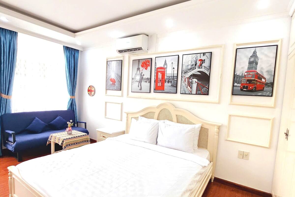 A hotel room with a large bed, a blue sofa, and a small table with a lamp. There are framed pictures on the wall and a window with curtains.