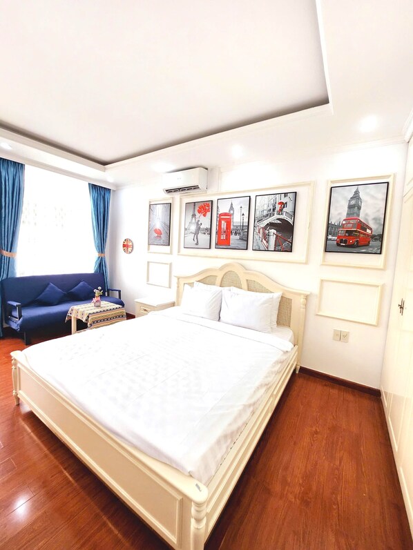 Habitación de hotel con una cama grande, un sofá azul y una mesita de noche con una lámpara. Hay cuadros en la pared y una ventana con cortinas.