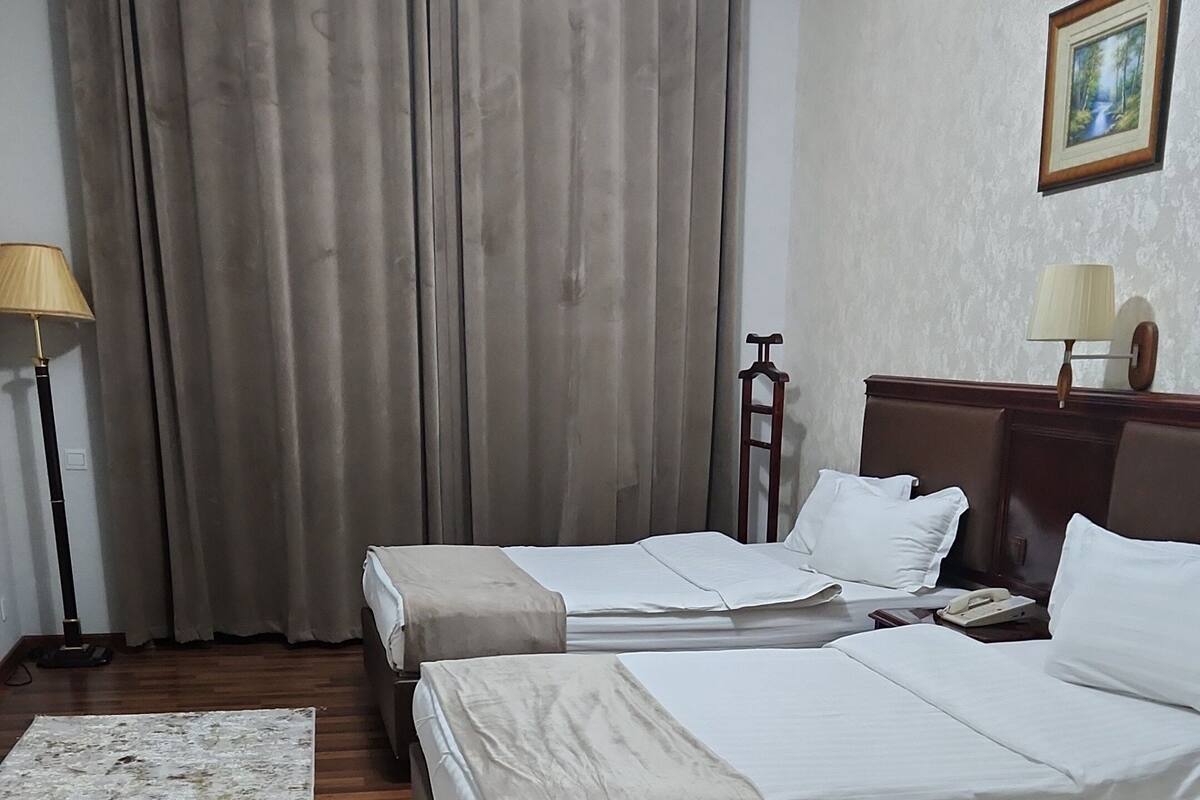 A hotel room with two beds, a wooden floor, a large window with curtains, a standing lamp, and a framed picture on the wall.
