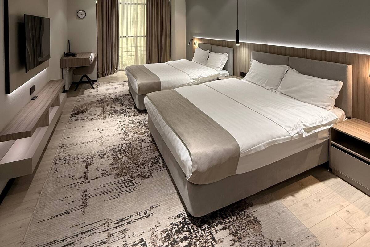 A modern hotel room with two beds, a flat-screen TV, a desk, and a large window with curtains.