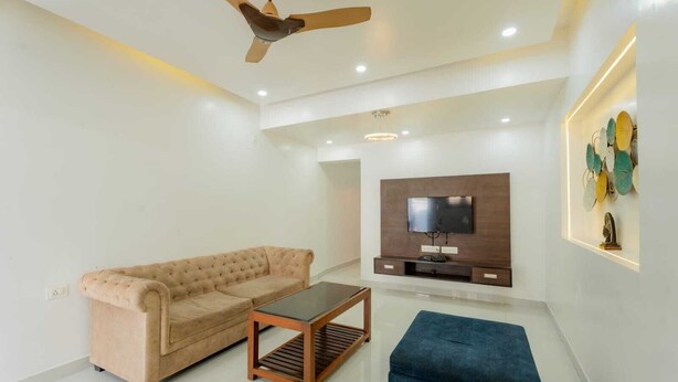 A modern living room with a beige sofa, a blue ottoman, a wooden coffee table, a ceiling fan, recessed lighting, a flat-screen TV mounted on a wooden entertainment unit, and a decorative wall piece. - New White Minar  (Calangute)