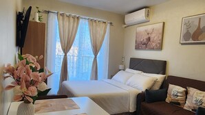 A hotel room with a bed, a sofa, a table with a vase of flowers, a TV, and a window with curtains.