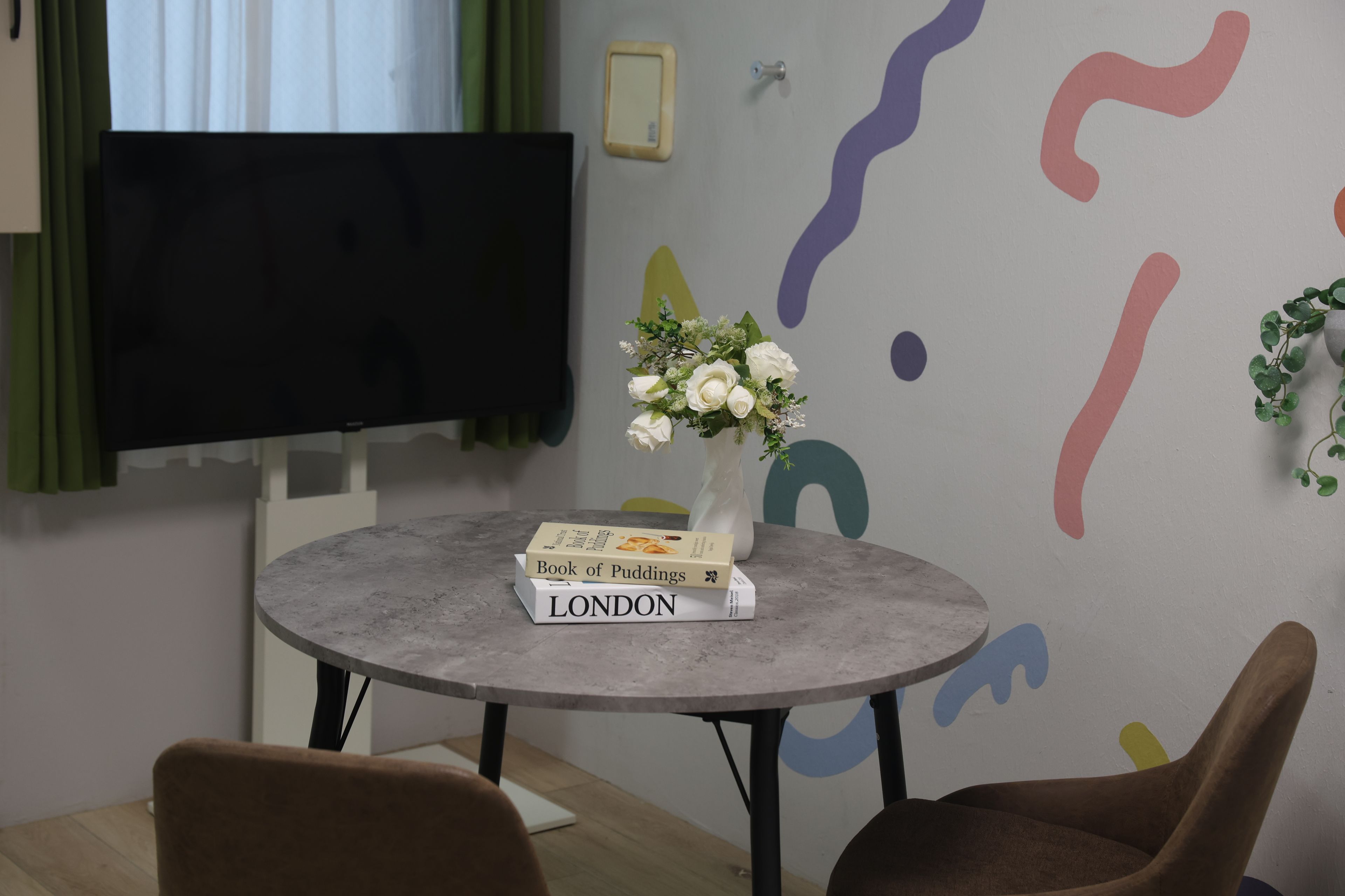 Una habitación con una mesa redonda, dos sillas, un televisor de pantalla plana, un florero con flores blancas y un libro titulado 'Book of Puddings LONDON'.