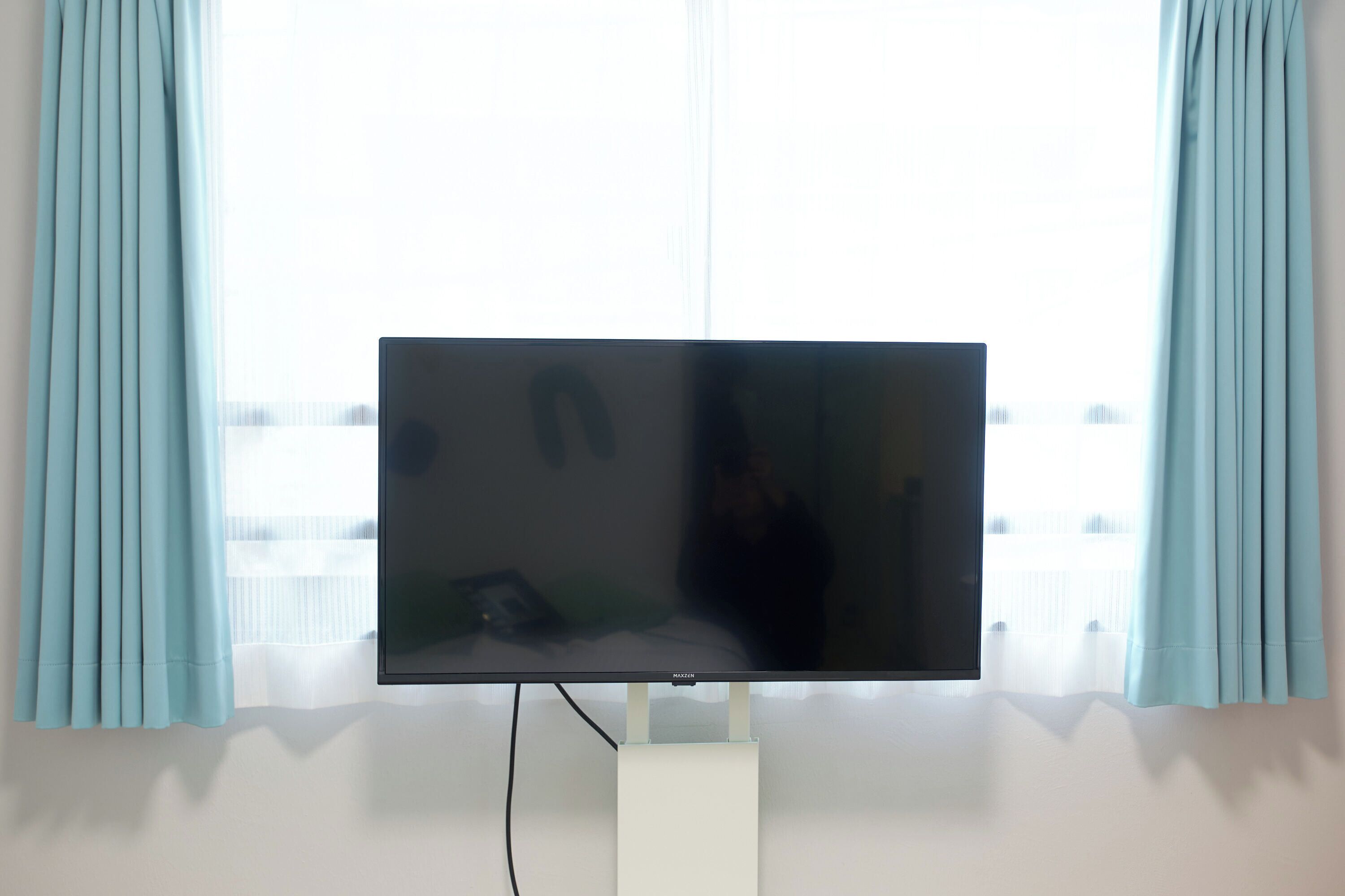 A flat-screen TV mounted on a stand in a room with light blue curtains and a window.