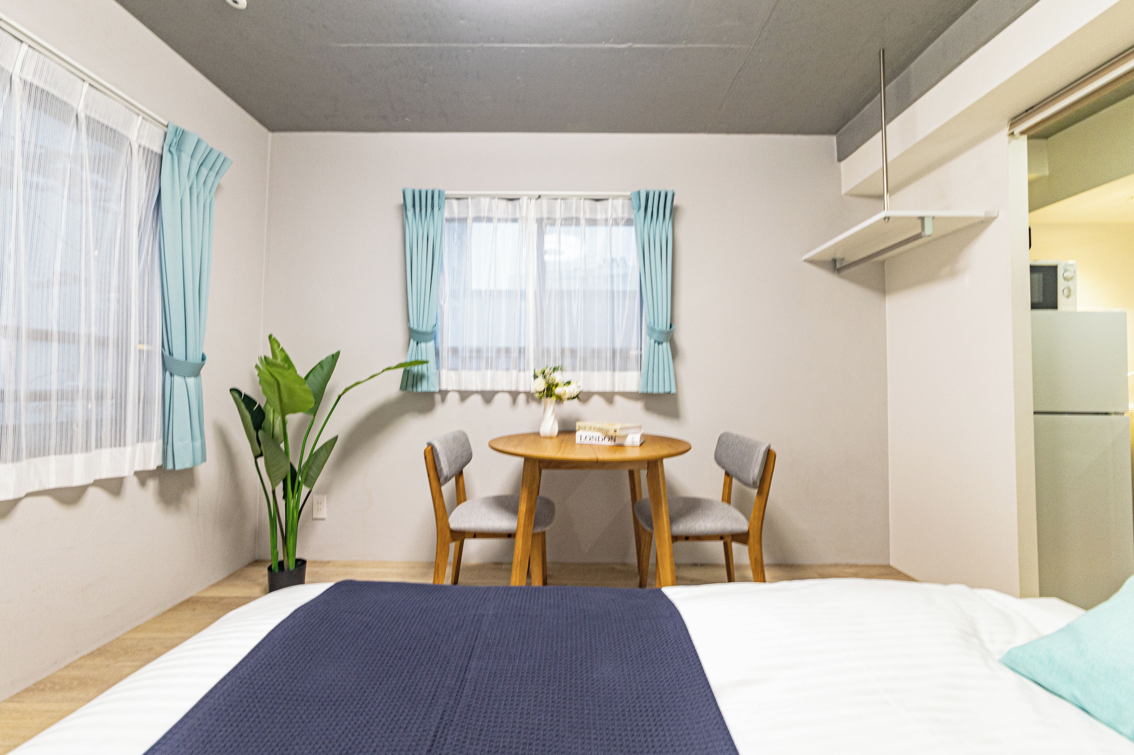 Habitación de hotel con cama, una mesa de comedor con dos sillas, una planta en maceta y una ventana con cortinas.