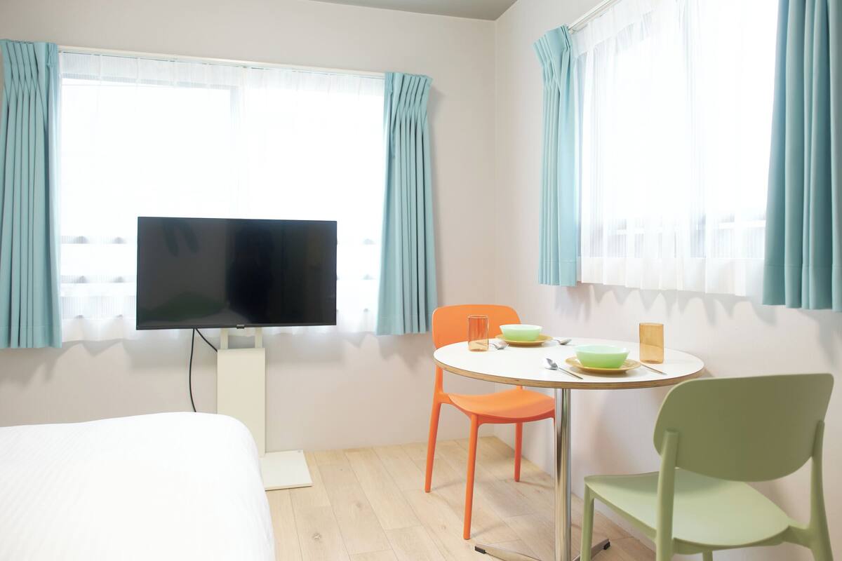 Une chambre d’hôtel comprenant un lit, une télévision, une table à manger avec des chaises et des fenêtres avec des rideaux.
