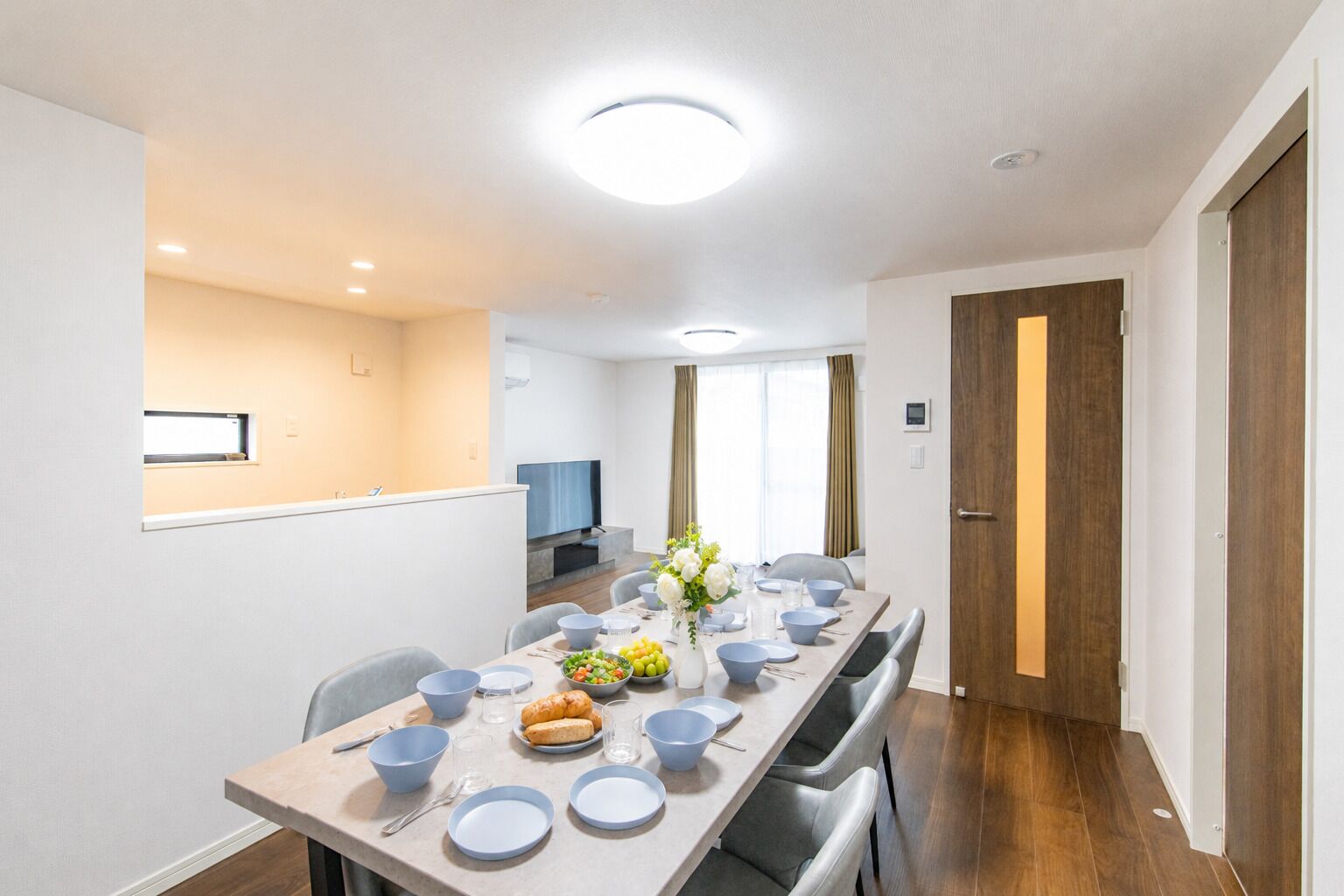 Un comedor moderno con una mesa larga dispuesta para cuatro personas, un televisor y una zona de cocina al fondo.