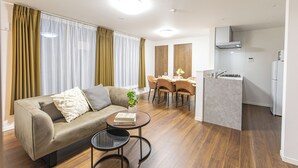 A modern living room with a sofa, a dining table, and chairs, a kitchen area with a stove and refrigerator, and a large window with curtains. - Ars Garden Omori (Tokyo)