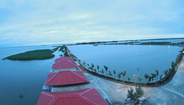 A resort with a long row of red-roofed buildings along a water body, surrounded by palm trees and a clear sky. - President Resort Kishoreganj (Mithamain)