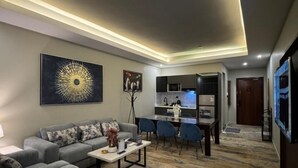 A modern living room with a grey sofa, a coffee table, and a dining area with blue chairs. - SWAT SERVICED APARTMENTS 5 (Al Khobar)