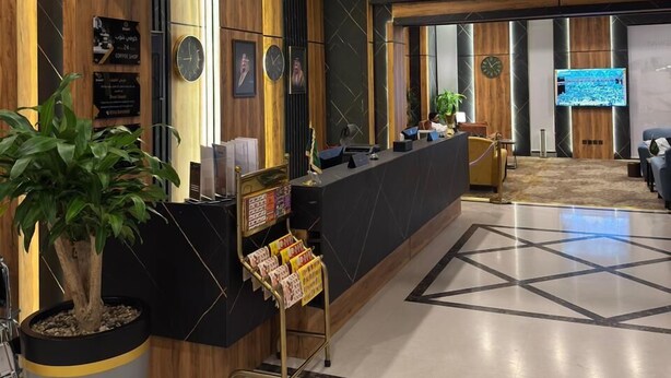 A reception area with a wooden reception desk, a potted plant, and a large screen displaying content. - SWAT SERVICED APARTMENTS 5 (Al Khobar)