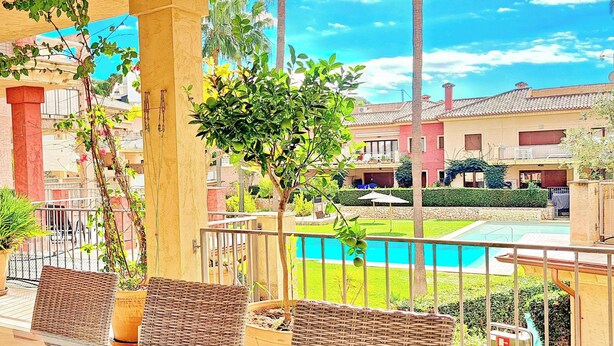 Un balcón con mobiliario de mimbre, plantas en macetas y vistas a una piscina y edificios.