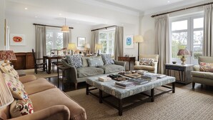A spacious living room with a large sofa, a coffee table with books, and a dining area with chairs and a table. - Elegant dog friendly home with panoramic views - The Old Vicarage (Aldsworth)