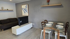 Ein modernes Wohnzimmer mit einem Glastisch, weißen Stühlen, einem dunklen Sofa, einem Flachbildfernseher auf einem weißen Schrank und einem Schmetterlingsbild an der Wand.