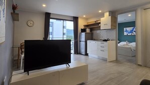 Eine moderne Wohnung mit Flachbildfernseher auf einer weißen Kommode, einer Küche mit weißen Schränken und einer Mikrowelle, einem Essbereich mit Tisch und Stühlen sowie einem Schlafzimmer mit einem Bett und einem blauen, abstrakten Gemälde an der Wand.