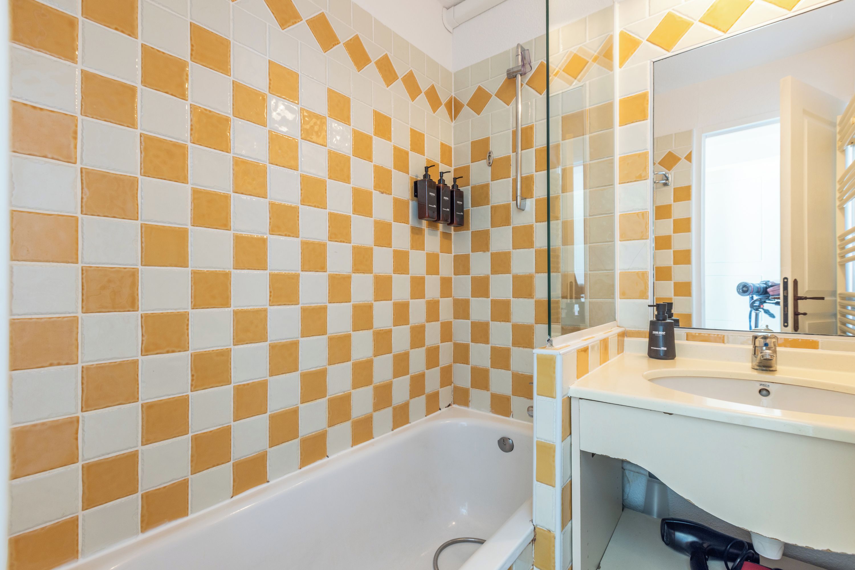 A bathroom with a checkered tile wall, a white bathtub, a sink with a mirror, and a showerhead.