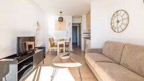 A modern living room with a flat-screen TV, a beige sofa, a wooden coffee table, and a dining area in the background. - Bright 1-bedroom sea & mountain view min. from beach (Cannes)