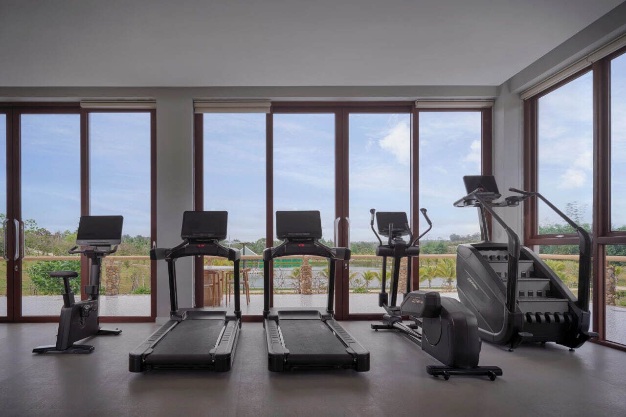 A gym with treadmills, elliptical machines, and a stationary bike, large windows with a view of greenery and a body of water.