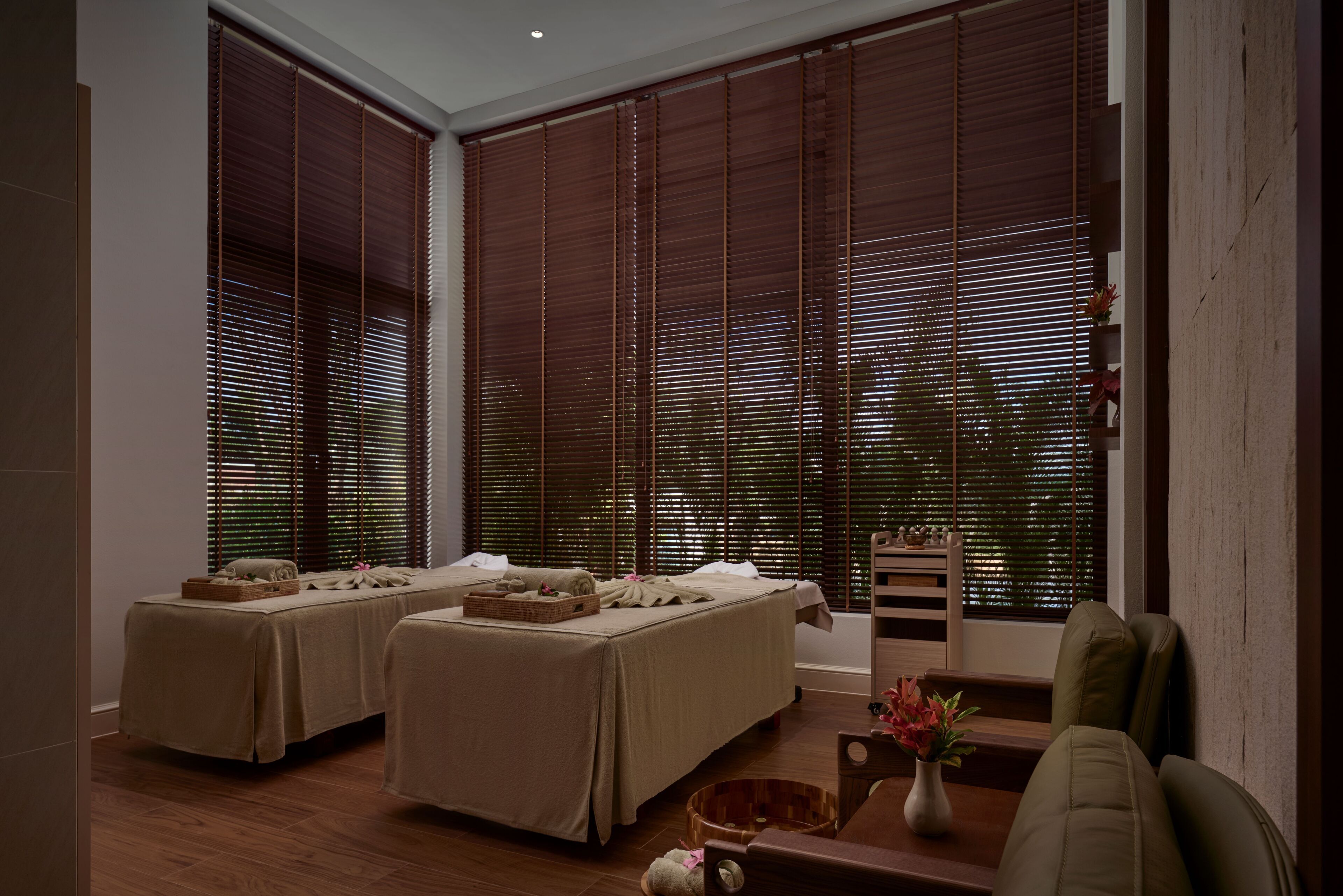 Une salle de spa équipée de deux tables de massage, de stores en bois, d’un parquet en bois et d’une petite plante posée sur une table.