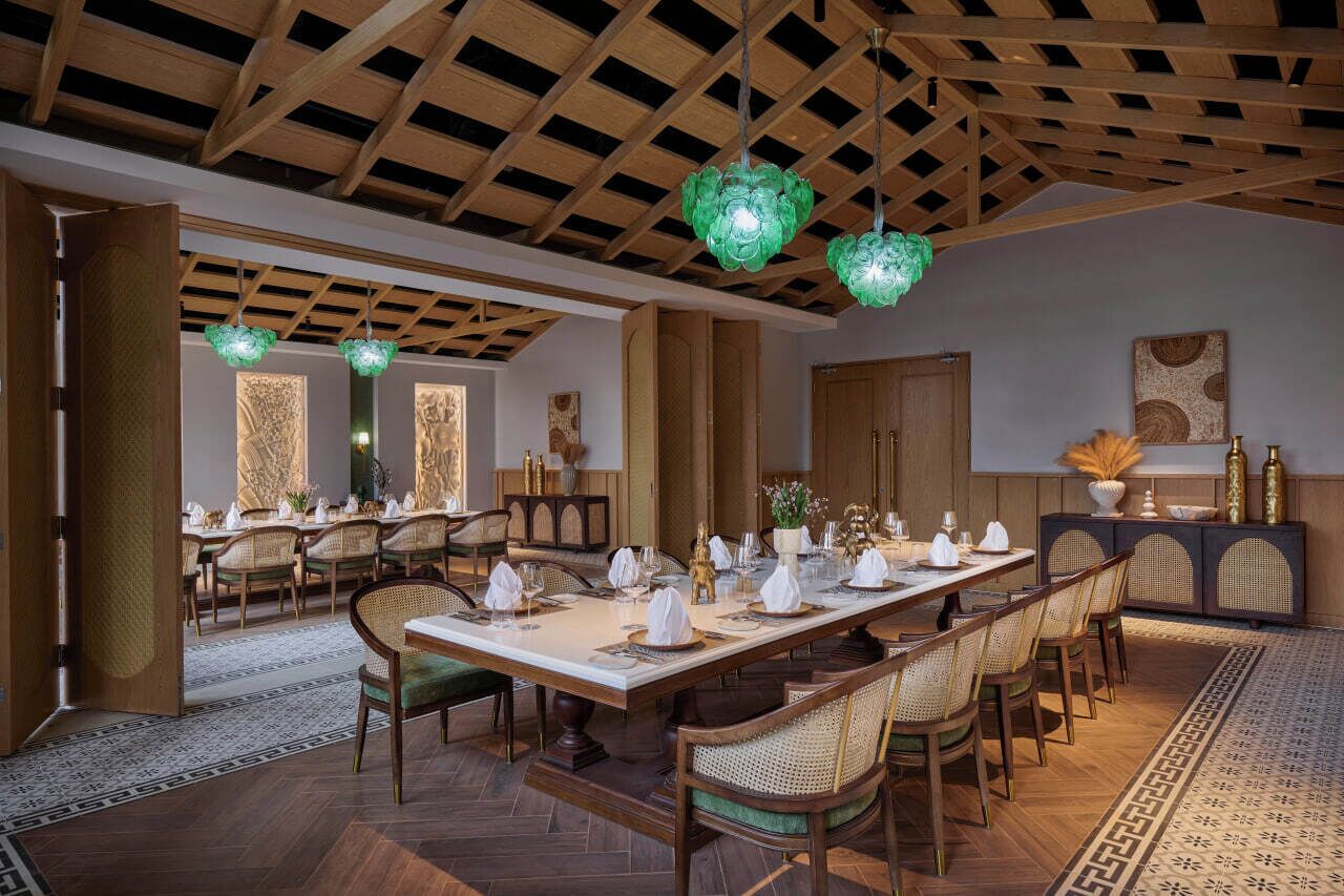 A dining area with a long table set for a meal, wooden ceiling beams, and green hanging light fixtures.