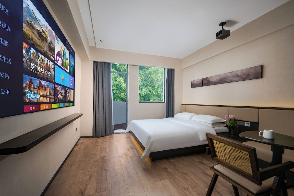 A modern hotel room with a large TV displaying various channels, a bed with white linens, a small table with a cup, and a view of greenery through the window.