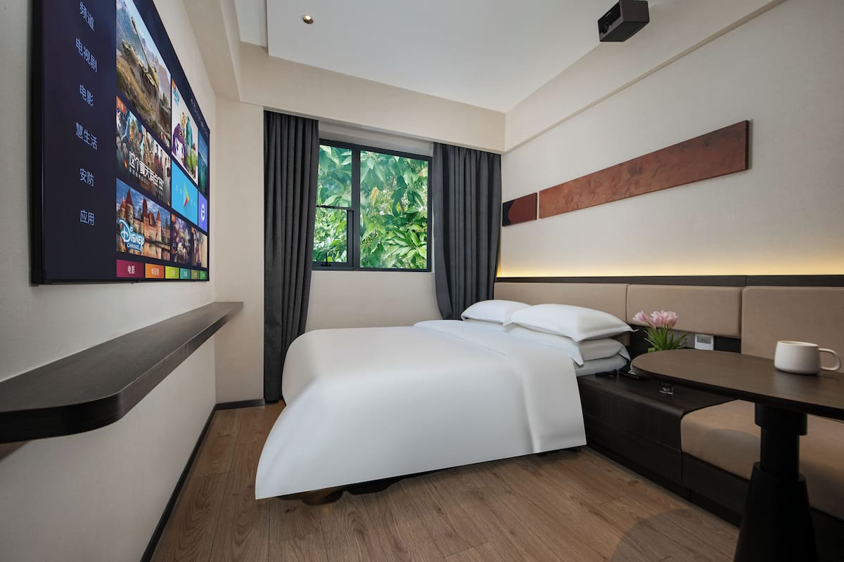 A modern hotel room with a large bed, a desk, a chair, a TV mounted on the wall, and a window with curtains.