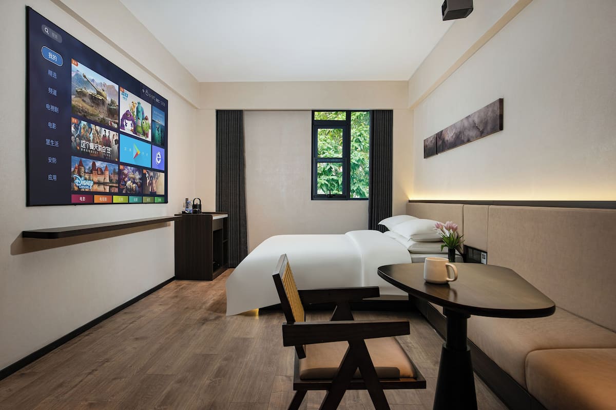 A modern hotel room with a large digital screen displaying various apps, a bed with white linens, a small table with a cup, a sofa, and a window with a view of greenery.