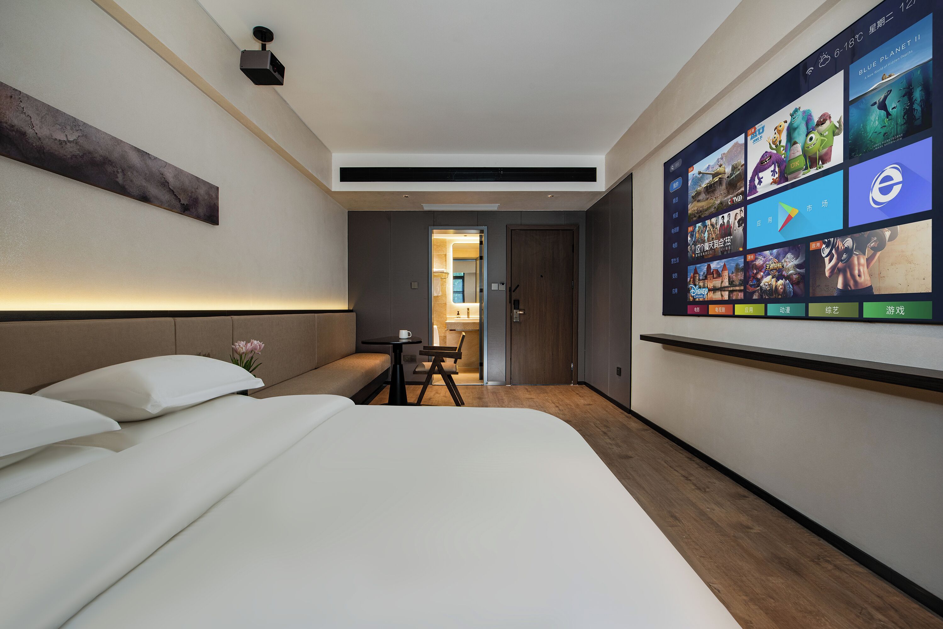 Una habitación de hotel moderna con una cama grande, un escritorio pequeño y un televisor montado en la pared mostrando diversos contenidos.