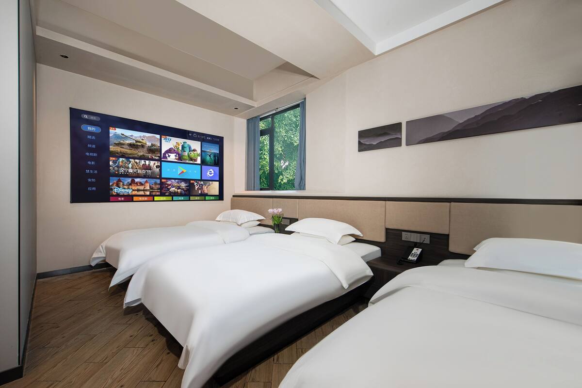 A hotel room with two beds, a large wall-mounted TV displaying various apps, a window with a view of trees, and a small vase with flowers on the nightstand.