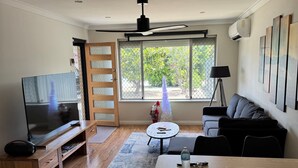 A modern living room with a flat-screen TV, wooden furniture, a dining table, and large windows offering a view of greenery outside.