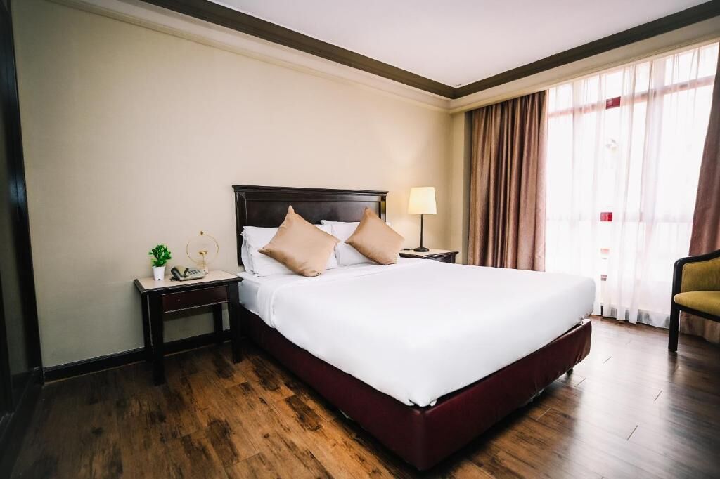 A hotel room with a large bed, a nightstand with a lamp, a small table with a plant, and a chair.