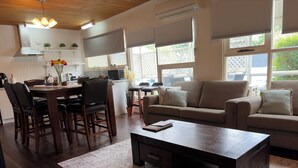 A modern living room with a dark wooden coffee table, beige sofa, and matching armchairs. A dining area with a wooden table and chairs is adjacent to the living room. Large windows with blinds provide natural light and views of the outdoors. - Hahndorf Retreat Cottage (Hahndorf)