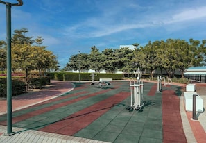 An outdoor fitness area with exercise equipment, a paved path, and greenery.