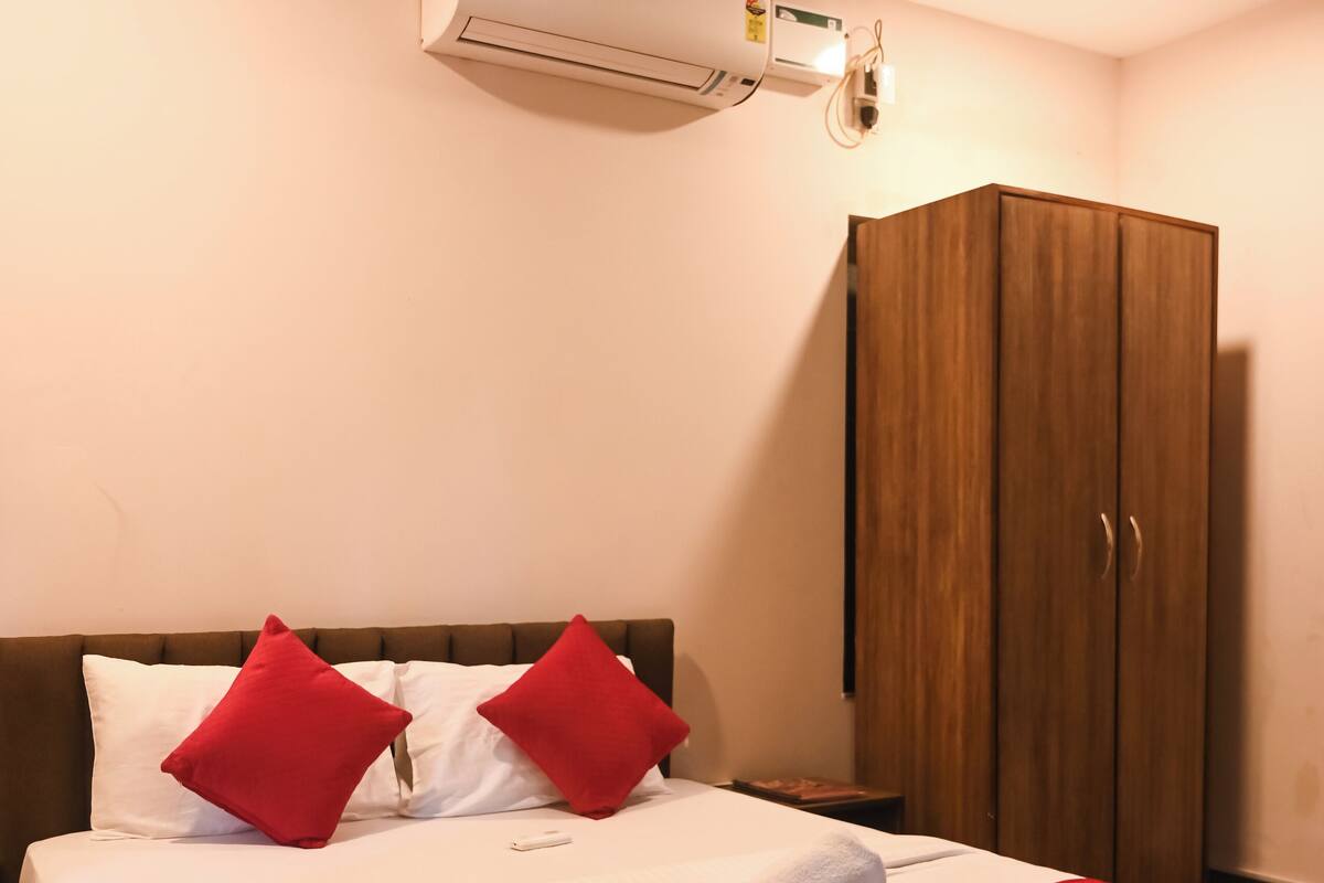A neatly made bed with red pillows, a wooden wardrobe, and an air conditioning unit above it.