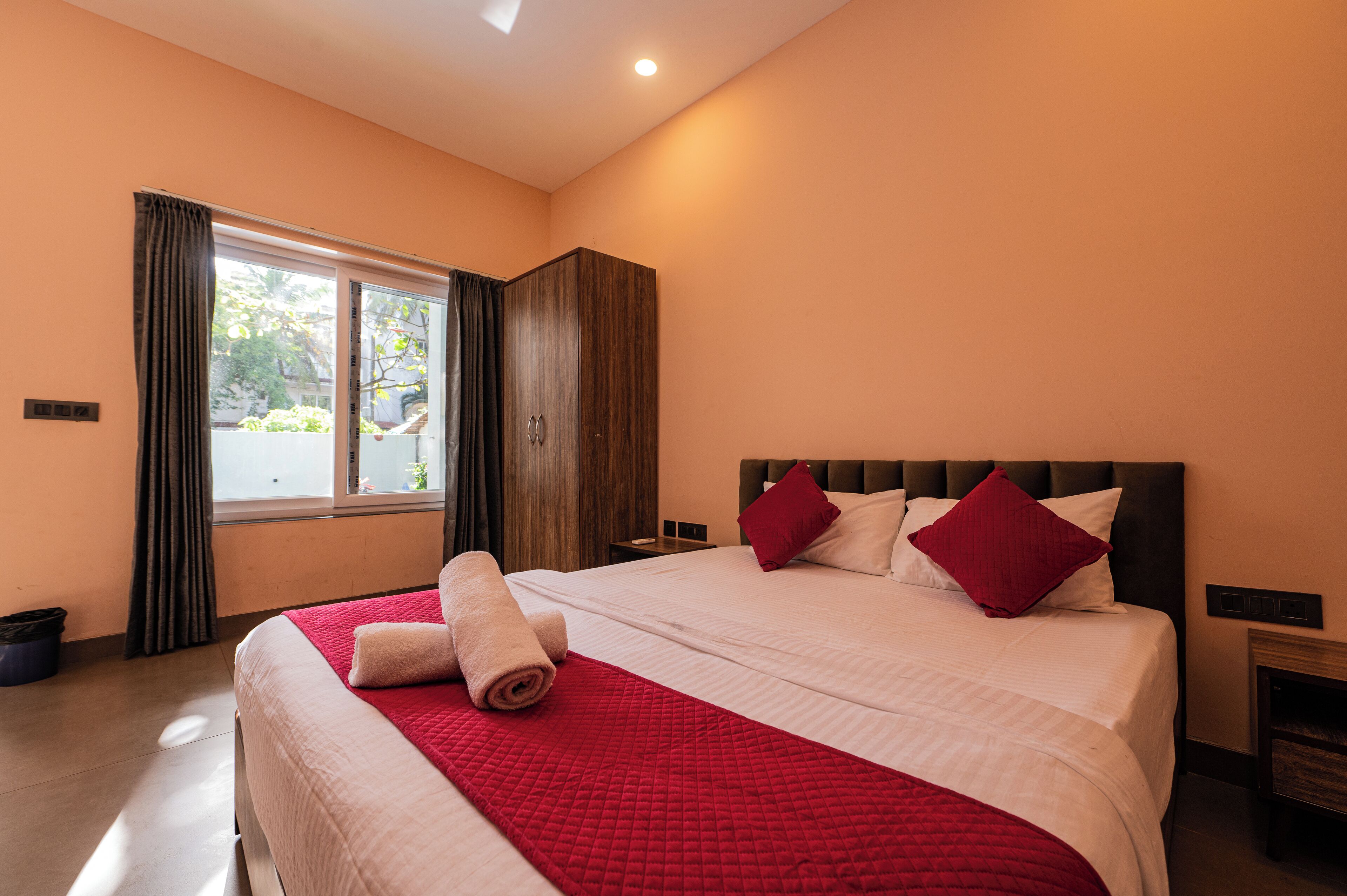 Una habitación de hotel con una cama grande, un armario, una ventana con cortinas y una mesita de noche.