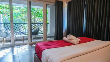 A hotel room with a bed, a red blanket, and a view of a balcony with chairs and greenery.
