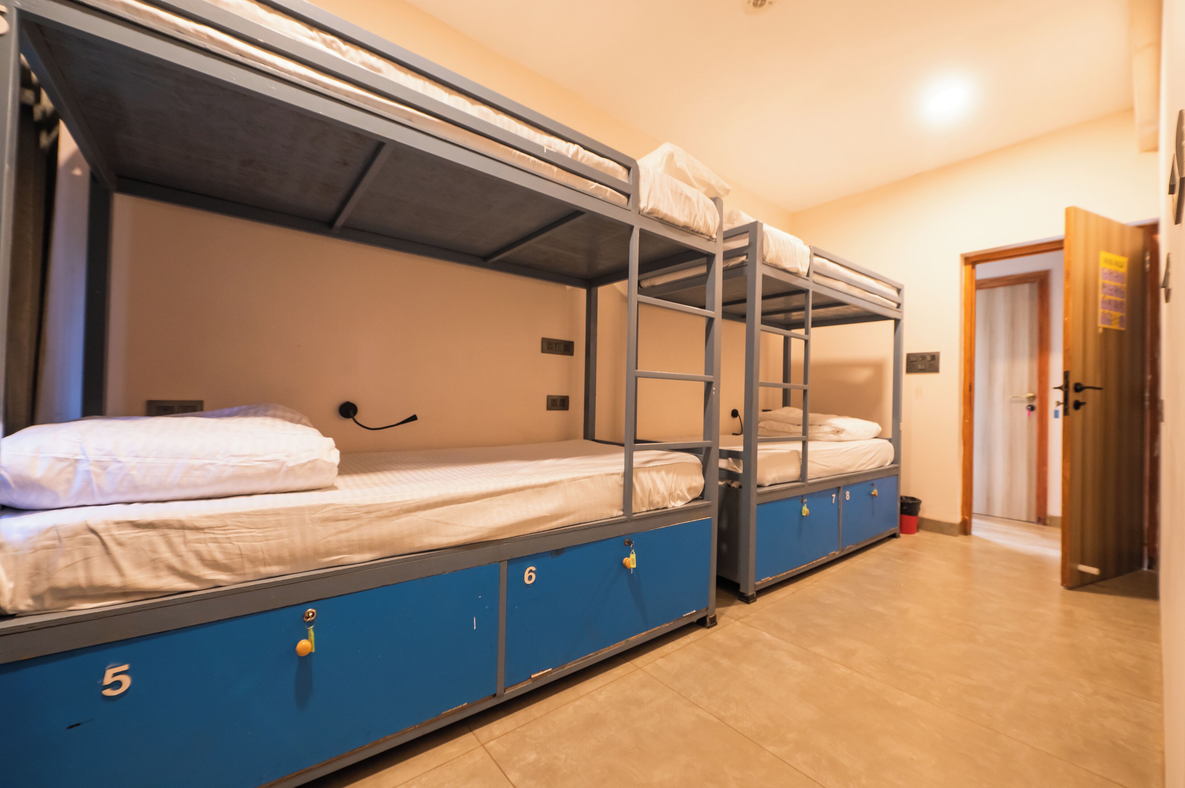 A dormitory room with bunk beds, numbered storage compartments, and a door leading to another room.