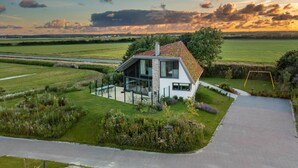 A modern house with a large glass front, surrounded by a well-maintained garden and a view of the countryside. - Modern, Luxurious, Sustainable Nature Villa With hot tub and Texel View (De Waal)