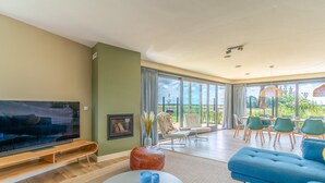 A modern living room with a large flat-screen TV, a blue sofa, and a dining area with green chairs. - Modern, Luxurious, Sustainable Nature Villa With hot tub and Texel View (De Waal)