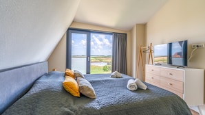 A bedroom with a large bed, a TV, a dresser, and a view of a landscape through a window. - Modern, Luxurious, Sustainable Nature Villa With hot tub and Texel View (De Waal)