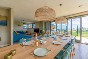 Uma sala de jantar com uma mesa de madeira comprida posta para seis pessoas, rodeada por cadeiras verdes. Há duas grandes luminárias pendentes de fibra acima da mesa.