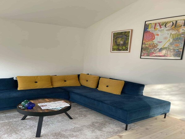 A modern living room with a blue sectional sofa, yellow cushions, a round coffee table with magazines, and two framed artworks on the wall. - 5 Star Holiday Home in Frederiksvaerk (Frederiksvaerk)