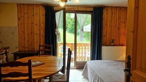 A room with a wooden floor, a round wooden table with chairs, a bed with a striped bedspread, and a view of a garden through a sliding glass door. - Apartment in Planay Near Hiking Trails (Beaufort)